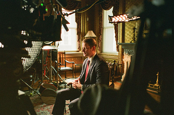 A man in a dark suit and red tie sits in a chair during a film production in a classic room.