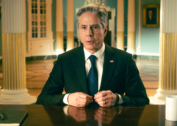 A man in a dark suit and blue tie sits at a polished desk in a room with large white columns.
