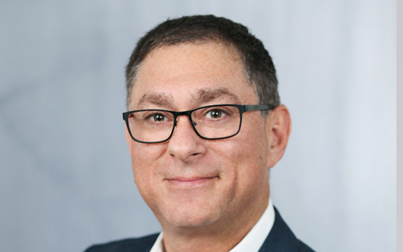 A man with short dark hair and black-rimmed glasses smiling slightly against a light blue background.