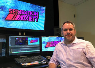 A man in a light pink checkered shirt sits before three computer monitors in a dark studio.