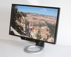 A black computer monitor on a silver ring stand displaying a scenic view of a canyon under a blue sky.