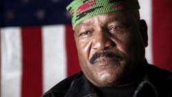 A man with a mustache wearing a green patterned cap in front of a blurred American flag.