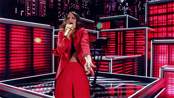 A woman in a red suit sings into a microphone on a stage with red neon lights.