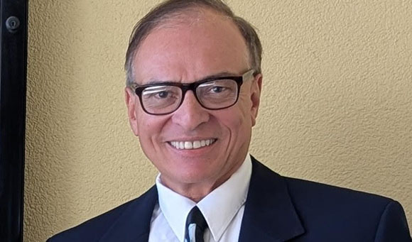 A smiling man with glasses wearing a dark suit and white shirt against a tan wall.