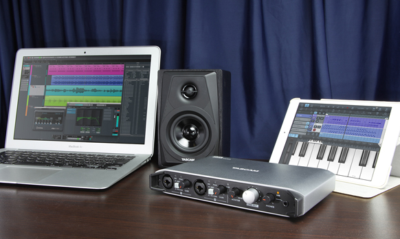 Laptop showing audio software, a black studio monitor, a silver audio interface, and a tablet with a piano keyboard.