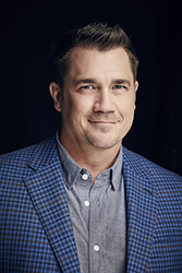 A man with short brown hair wearing a blue checkered blazer and a grey button-down shirt.