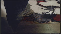 A person wearing dark pants and a tan shoe steps near a dark liquid spill on a light-colored floor.
