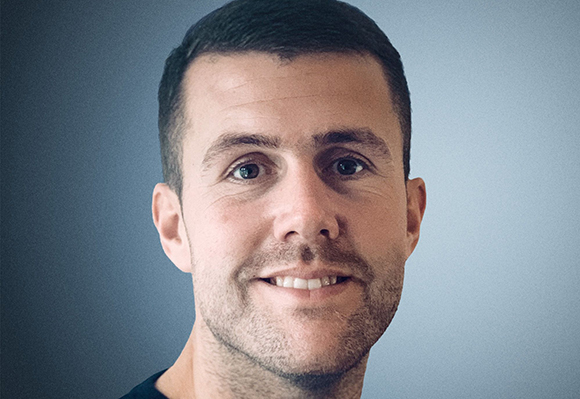 Close-up portrait of a man with short dark hair and light stubble against a gray background.