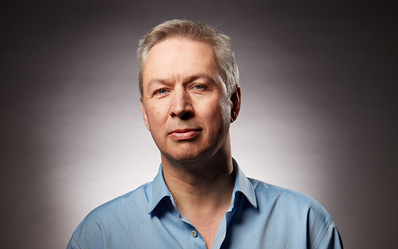 A middle-aged man with short graying hair wearing a light blue button-down shirt against a gray background.