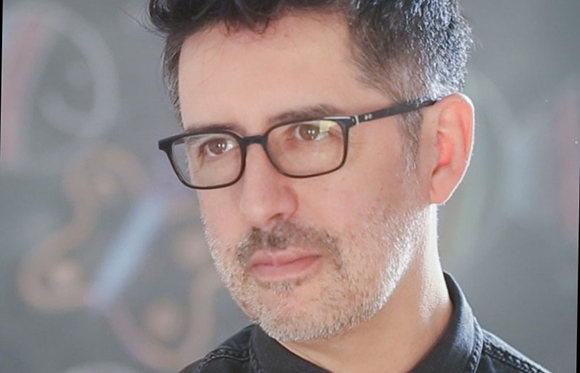 A man with dark hair, glasses, and light facial stubble looking slightly off-camera.