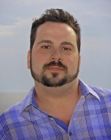 A man with dark hair and a groomed beard wearing a blue and white plaid button-down shirt.
