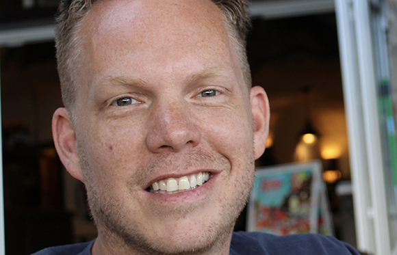 Close-up of a smiling man with light skin and short blonde hair looking toward the camera.
