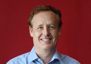 A smiling man with reddish-brown hair wearing a light blue button-down shirt against a solid red background.