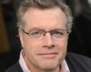 Close-up portrait of a man with light hair and glasses wearing a pink shirt and dark sweater.
