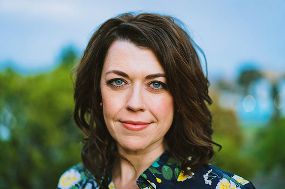 A woman with wavy brown hair and blue eyes looks directly at the camera against a blurred outdoor background.
