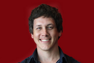 A man with wavy brown hair smiling at the camera against a solid red background.