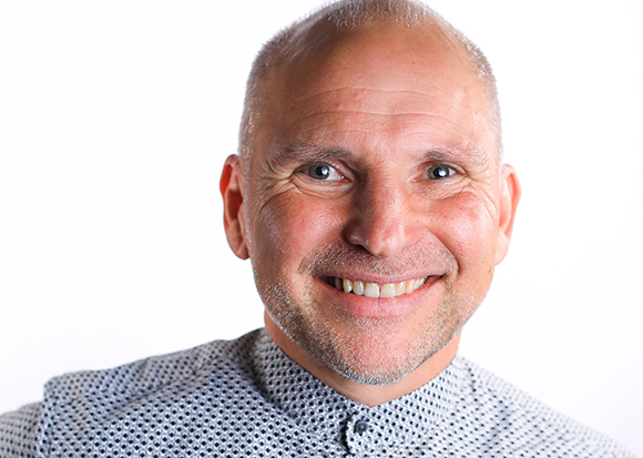 A smiling man with a shaved head and light stubble wearing a patterned grey shirt against a white background.