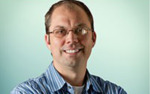 A man with short brown hair and glasses smiling against a light blue background.