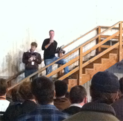 Two men stand on a wooden staircase addressing a seated audience in a large room.