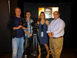 Four people stand behind a camera on a tripod in a studio setting. A screen is visible behind them.