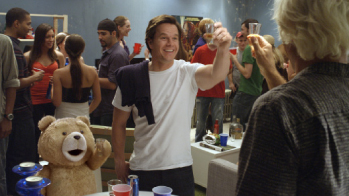 A man in a white t-shirt raises a shot glass to toast a group of people at a crowded indoor party.