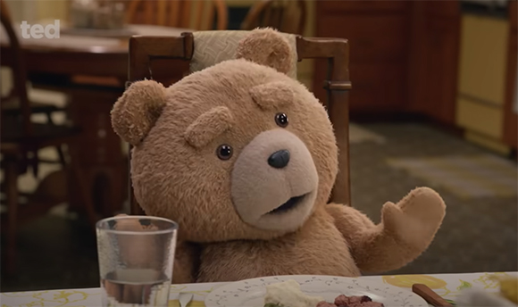 A brown teddy bear sits at a dining table with a glass of water and food in front of it.