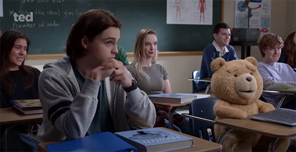 Students sit at desks in a classroom while a large teddy bear sits at a desk in the foreground.