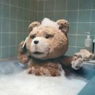 A brown teddy bear sitting in a bathtub filled with white soap bubbles against a tiled wall.