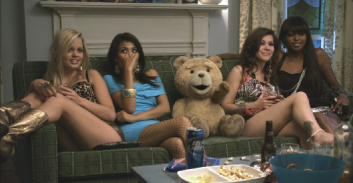 Four women sit on a green couch next to a large teddy bear in a living room.