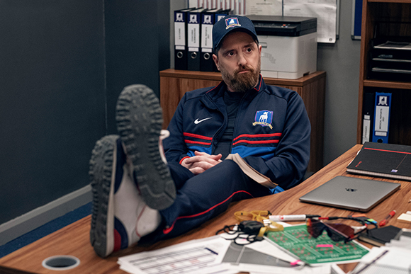 A bearded man in a blue tracksuit and cap sits at a desk with his feet resting on the table.
