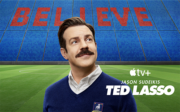 Jason Sudeikis smiling in front of a blue stadium screen displaying the word BELIEVE in red.