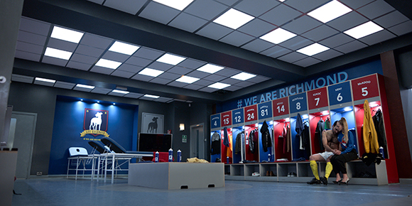 A locker room with blue walls, rows of numbered lockers, and two people sitting on a bench.