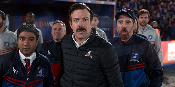 A group of men wearing blue and black athletic jackets with crests, looking surprised in a stadium setting.