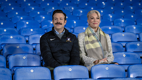 A man with a mustache and a blonde woman sit side-by-side in blue stadium seating.