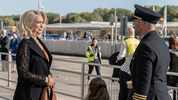 A blonde woman in a black lace coat talks to a man in a dark pilot uniform outdoors.