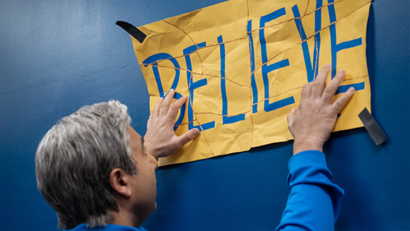 A man in a blue shirt tapes a torn yellow paper sign that says BELIEVE to a dark blue wall.