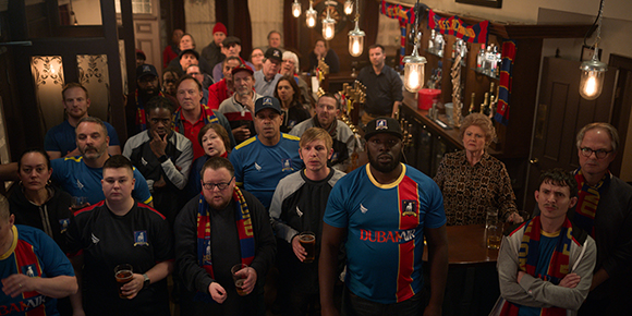 A group of people gathered in a dimly lit pub, many wearing blue and red football jerseys and scarves.
