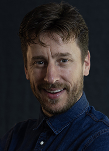 A man with brown hair and a beard smiling against a dark background.