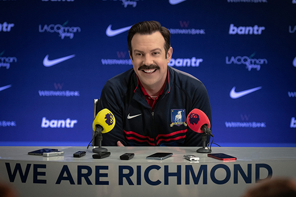 A man with a mustache and a navy blue jacket sits behind a table with microphones and a sign reading 'WE ARE RICHMOND'.