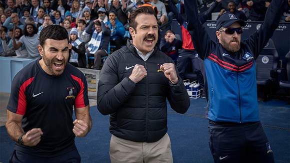 Three men in athletic gear cheering with clenched fists in a crowded stadium.