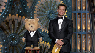 Mark Wahlberg in a tuxedo standing next to a teddy bear wearing a tuxedo on a stool.