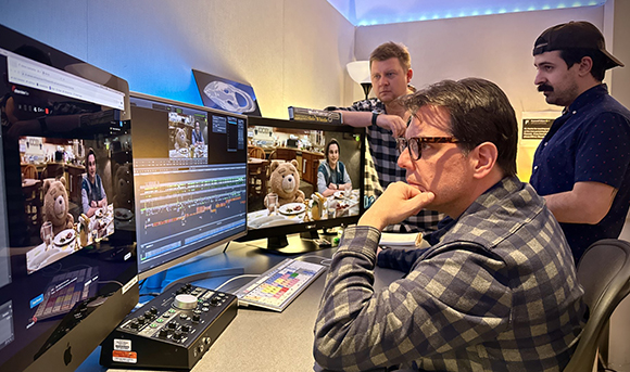 Three men sit at a desk with multiple computer monitors and editing equipment in a studio.