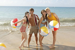 Four young adults standing on a beach holding colorful beach balls near the ocean waves.