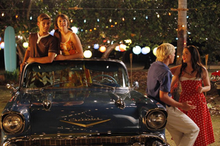 Four young people socialize around a vintage blue car under string lights at night.