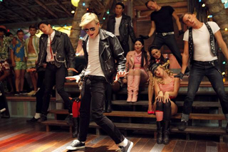 A group of young people in leather jackets and casual wear dancing in a dimly lit room with wooden floors.