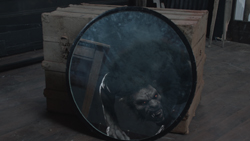 A circular mirror reflecting a dark, monstrous face with glowing red eyes in front of wooden crates.