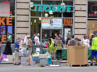 People standing near boxes and electronic equipment on a sidewalk in front of a Tekserve storefront.