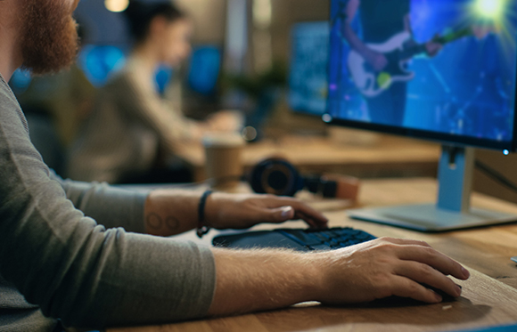 A person's hands typing on a keyboard and using a mouse at a wooden desk in a workspace.
