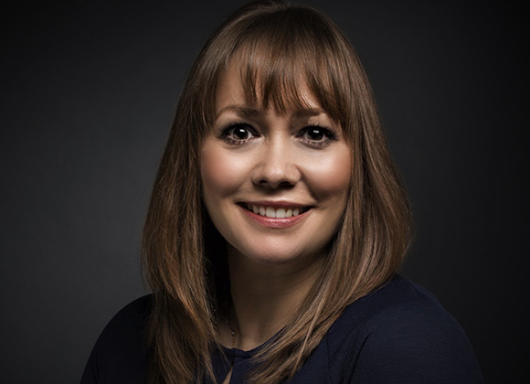A smiling woman with shoulder-length brown hair and bangs against a dark background.