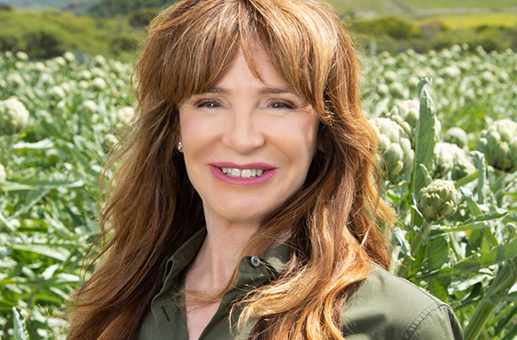 A woman with long wavy reddish-brown hair smiling in a field of green artichoke plants.